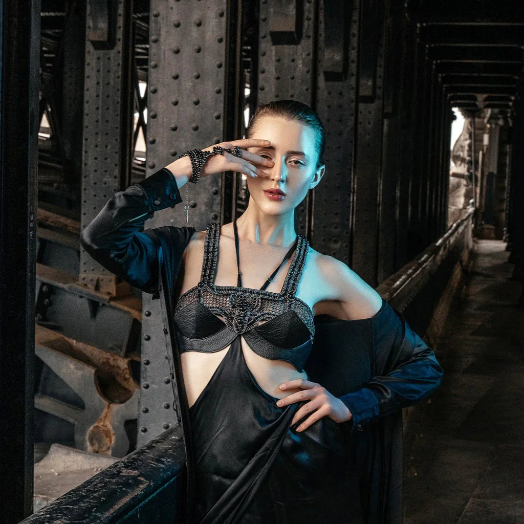 Fair skinned model wearing beaded black top and hand piece by Object & Dawn in a black silk dress standing in a dark, industrial setting